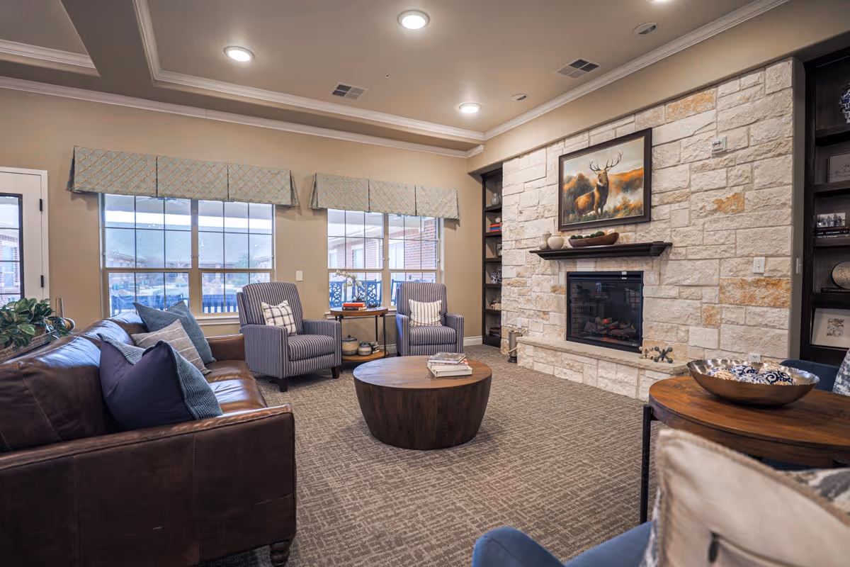 A cozy living room with a stone fireplace featuring a painting of a deer above it. The room has a brown leather sofa with blue and gray pillows, two striped armchairs, a round wooden coffee table with books on it, and built-in shelves on either side of the fireplace. Large windows with patterned valances let in natural light.