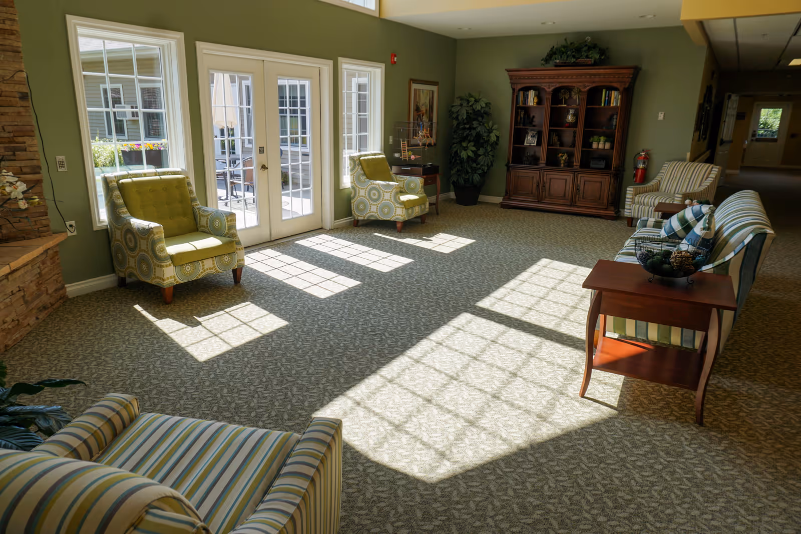 A bright and spacious living room area with large windows and glass doors letting in sunlight. The room features patterned armchairs and a striped sofa arranged around a wooden side table with a decorative bowl. A wooden bookshelf with plants and books is against the far wall, and a potted plant is in the corner. The walls are painted green and the carpet has a subtle leaf pattern.