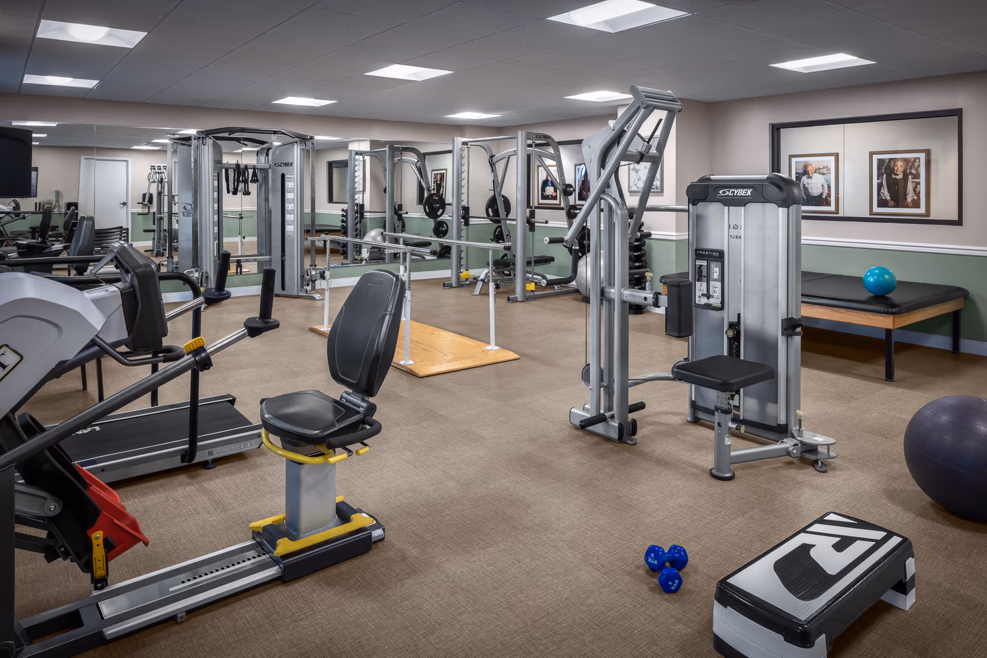 A well-equipped fitness room in a senior living facility featuring various exercise machines including a recumbent bike, weight machines, a treadmill, free weights, and a step platform. The room has beige carpet, light green and white walls, framed pictures, and large mirrors along one wall.
