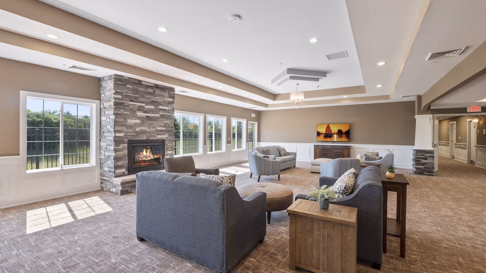 A spacious and well-lit living room area in an assisted living facility featuring multiple gray upholstered sofas and armchairs arranged around a round ottoman. A modern stone fireplace with a fire burning is on the left wall beneath large windows letting in natural light. The walls are painted beige with white wainscoting, and a chandelier hangs from the tray ceiling. A colorful painting is mounted on the far wall above a wooden cabinet. The carpet is light brown, and there is a hallway visible to the right.