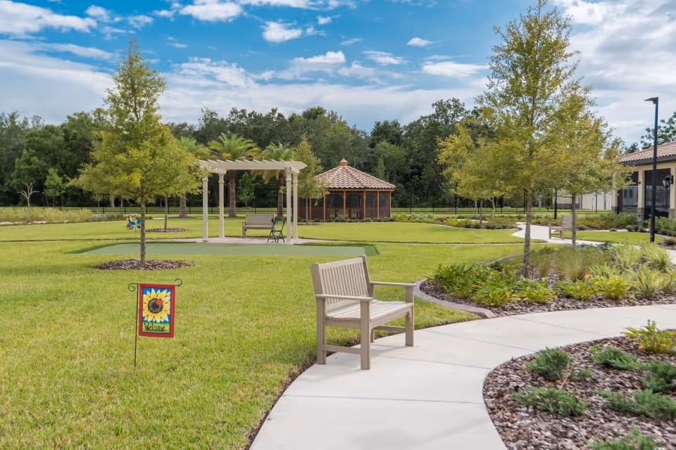 A well-maintained outdoor garden area with a curved concrete pathway, green grass, small trees, and landscaped plants. There is a wooden bench along the pathway and a small decorative sign with a sunflower and the word 'Welcome'. In the background, there is a white pergola with seating underneath and a gazebo with a tiled roof surrounded by trees and greenery.