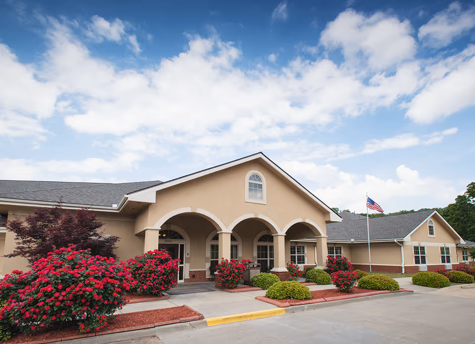 Exterior view of Covington Court Health and Rehabilitation Center showing a beige building with arched entrance, well-maintained landscaping with red flowers and green bushes, and an American flag on a flagpole under a partly cloudy blue sky.