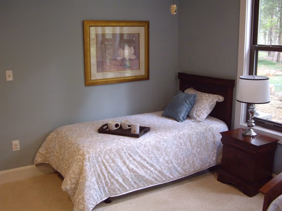 A single bed with a patterned beige and white bedspread and two pillows, one beige and one blue, is positioned against a light blue wall. A wooden nightstand with a silver lamp and white lampshade is next to the bed by a window showing an outdoor view. A framed artwork hangs above the bed. A tray with two white mugs and a small container is placed on the bed.
