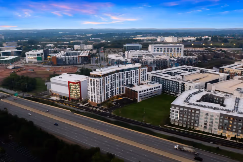 Aerial view of a large residential and commercial complex near a highway at dusk, with multiple modern buildings and a partly cloudy sky with a colorful sunset.