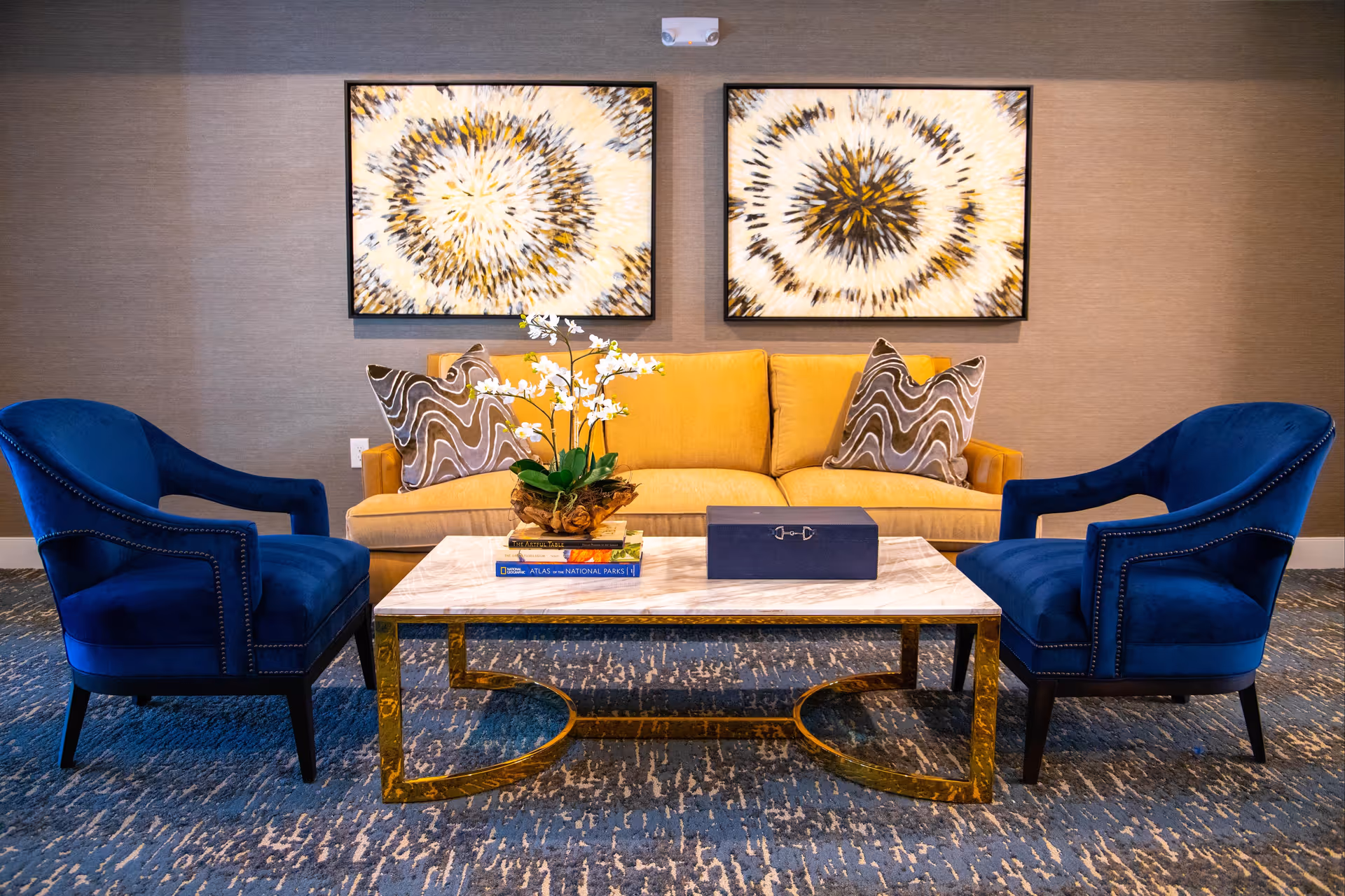 A stylish living room area featuring a mustard yellow sofa with two patterned pillows, flanked by two deep blue upholstered armchairs. In front of the seating is a white marble coffee table with a gold metal base, decorated with a small plant and a couple of books. The wall behind the sofa has two abstract circular art pieces in shades of white, black, and gold.