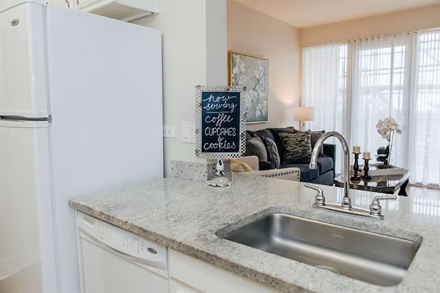 View of a modern kitchen countertop with a stainless steel sink and faucet, a white refrigerator, and a dishwasher. A small decorative sign on the counter reads 'now serving coffee cupcakes & cookies.' In the background, there is a living room area with a dark sofa, a side table with candles, a lamp, and large windows with sheer curtains letting in natural light.