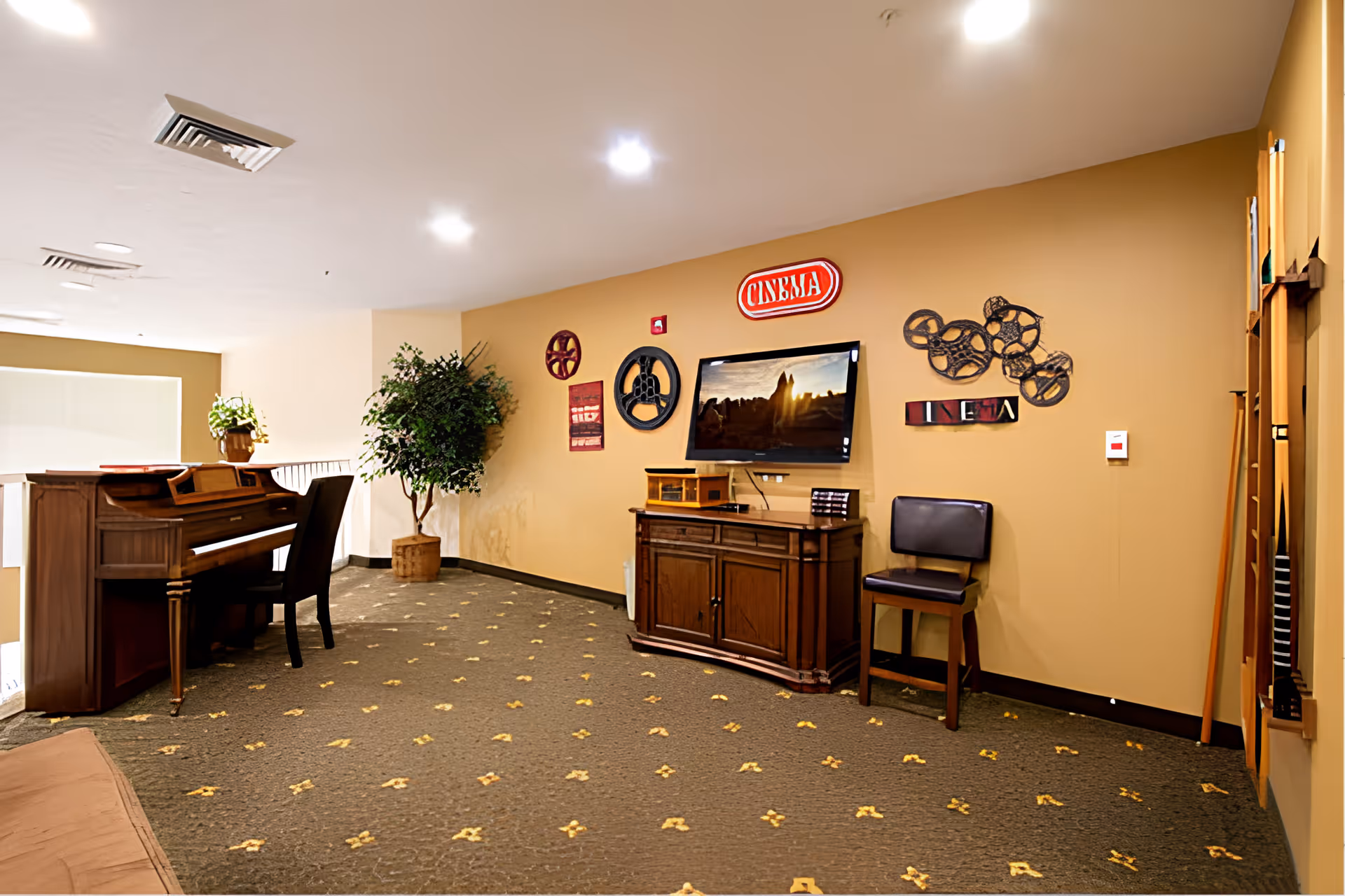 Interior common area with a piano and chair on the left, a potted plant in the corner, and a wall-mounted TV on the right. The wall behind the TV has decorative film reels and a red sign that says 'CINEMA'. There is a wooden cabinet below the TV and a single chair next to it. The carpet is patterned with small yellow designs.