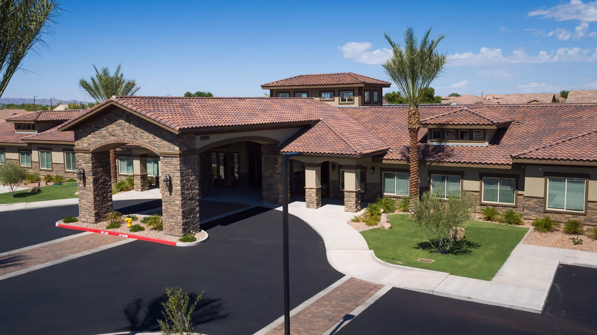 Exterior view of a single-story senior living facility with a stone and stucco facade, red tile roof, and a covered entrance. The building is surrounded by landscaped greenery including palm trees and bushes, with a paved driveway and sidewalks under a clear blue sky.