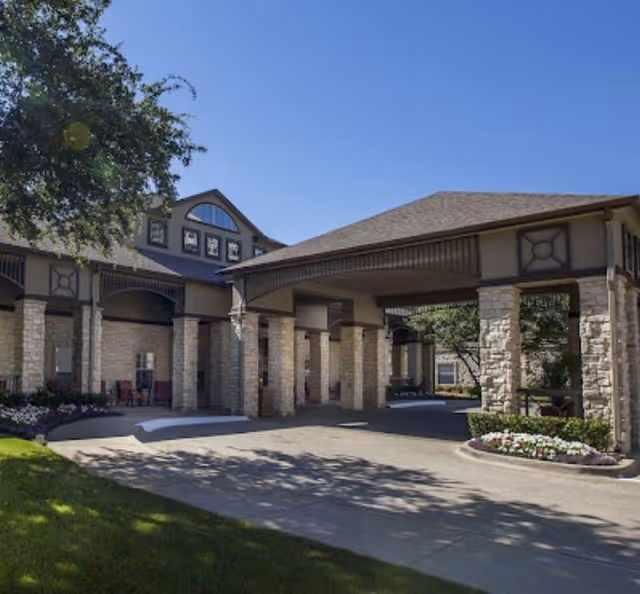 Exterior view of The Reserve at North Dallas Assisted & Senior Living facility showing a covered entrance with stone pillars, landscaped flower beds, and a clear blue sky.