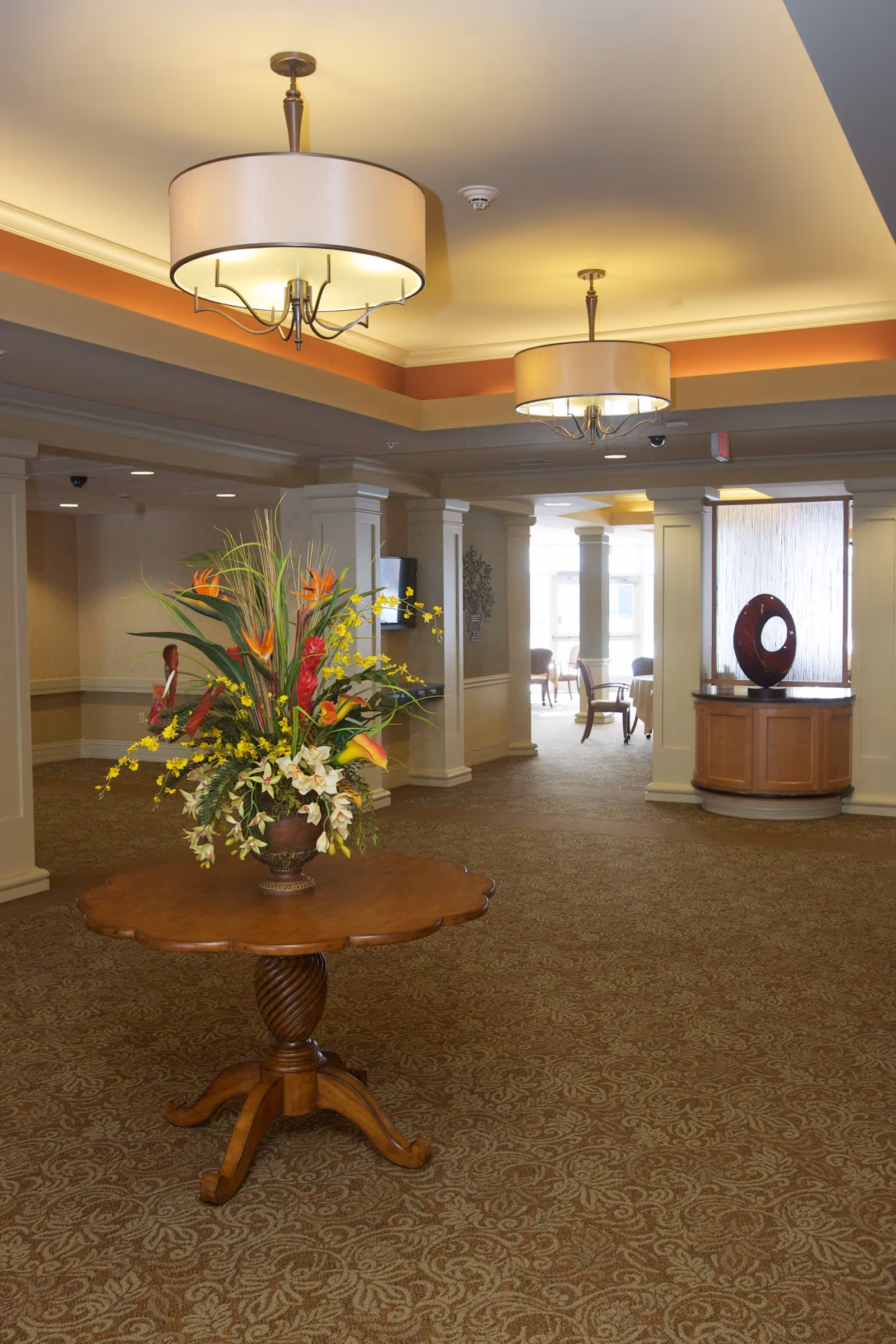 Interior view of a senior living facility lobby area with a round wooden table in the center holding a large floral arrangement. The space features patterned carpet, decorative columns, two large ceiling light fixtures, and a wooden cabinet with a modern sculpture in the background.