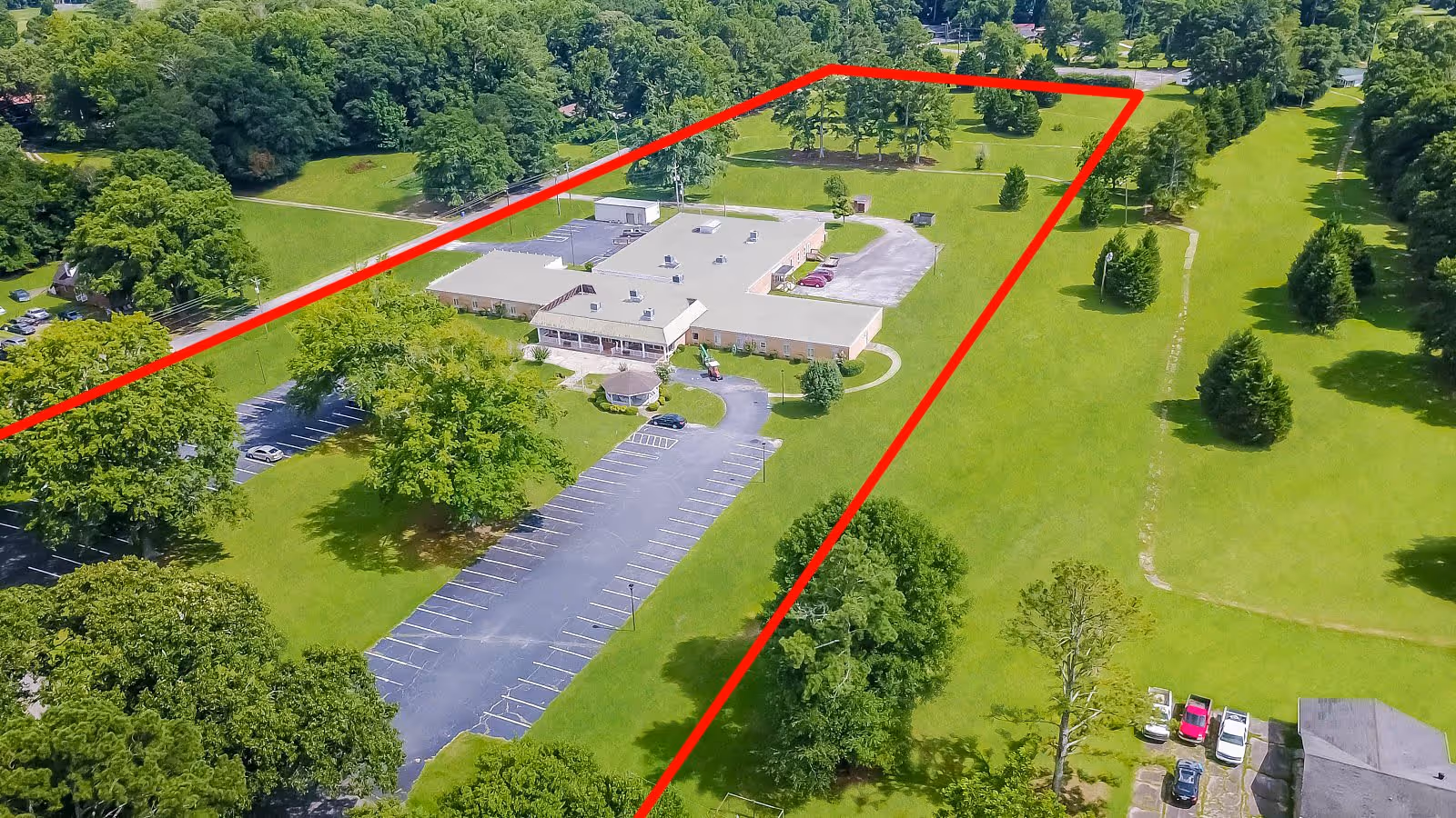 Aerial view of Front Porch of Bowdon Senior Living facility showing a single-story building surrounded by green lawns and trees. There is a large parking lot with marked spaces, a gazebo near the building, and several cars parked near the entrance. The property is outlined in red, highlighting the facility's boundaries within a lush, green landscape.