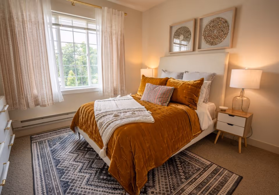 Cozy bedroom with a bed dressed in mustard-colored bedding, two bedside tables with lamps, a patterned rug, and a large window with flowing curtains.