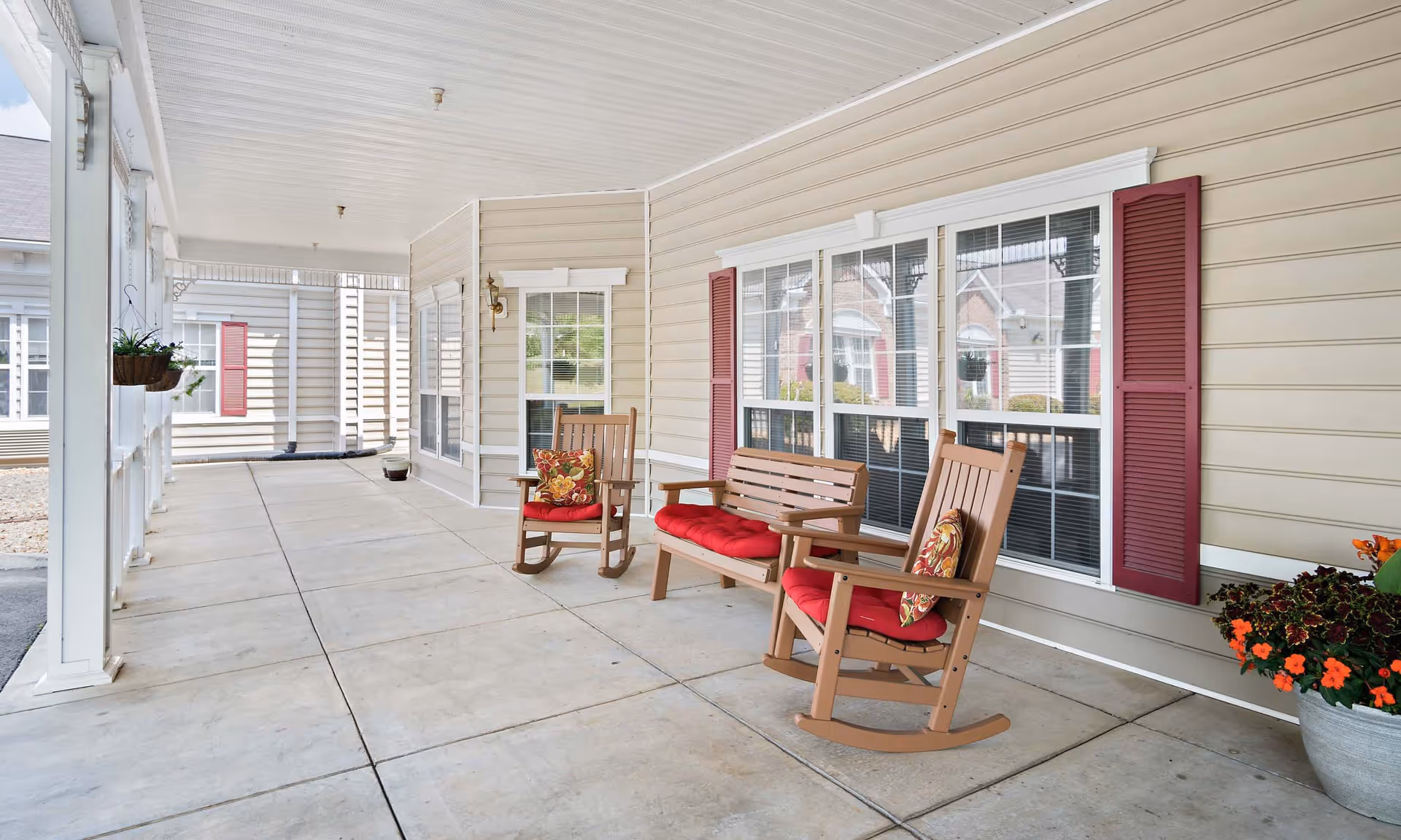 Covered outdoor porch area with beige siding walls and white trim, featuring two wooden rocking chairs and a wooden bench with red cushions and floral pillows. There are potted plants with flowers on the right side and hanging plants on the left. Large windows with red shutters are visible on the wall behind the seating.
