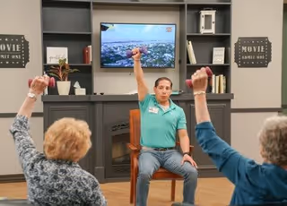 A fitness instructor leads an exercise session with two elderly women in a living room setting. The instructor, seated on a chair, raises one arm holding a small dumbbell, while the two women mirror the movement, each holding a dumbbell. Behind them is a wall-mounted TV and shelves with decorative items and books.