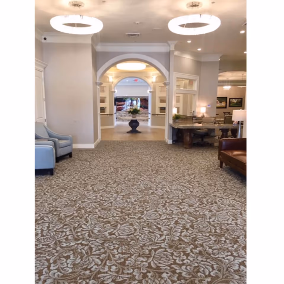 Interior view of a senior living facility hallway with patterned carpet, light gray walls, and modern circular ceiling lights. There are blue armchairs on the left, a brown leather sofa on the right, and a reception desk with chairs and lamps further back on the right. An arched doorway leads to another area with a large decorative vase and a scenic wall mural in the background.