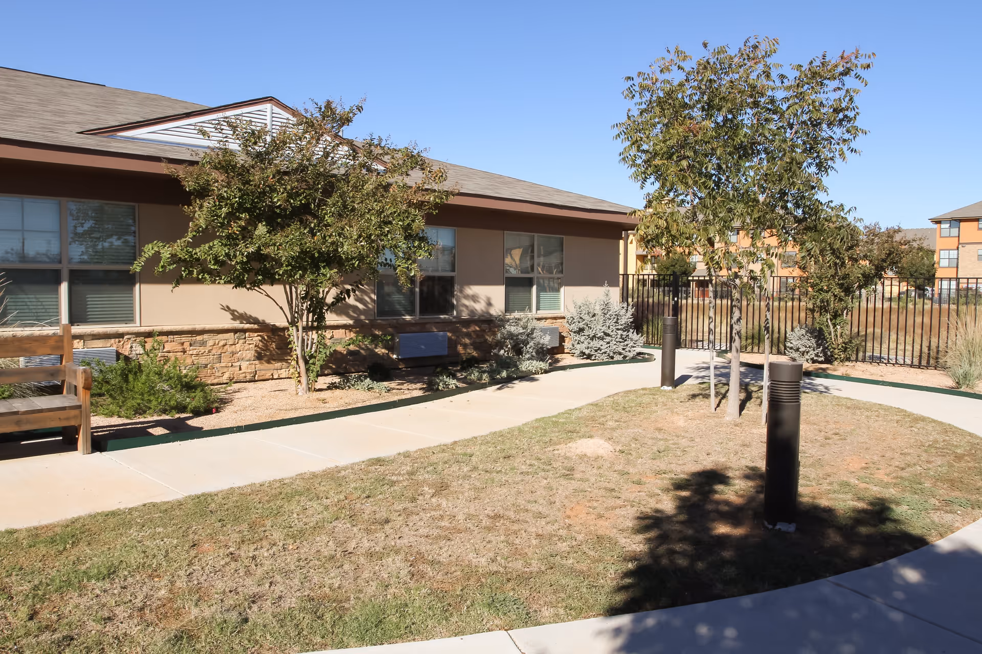 Outdoor courtyard area at The Courtyards Assisted Living and Memory Care featuring a paved walkway, small trees, shrubs, a wooden bench, and a building with windows in the background under a clear blue sky.