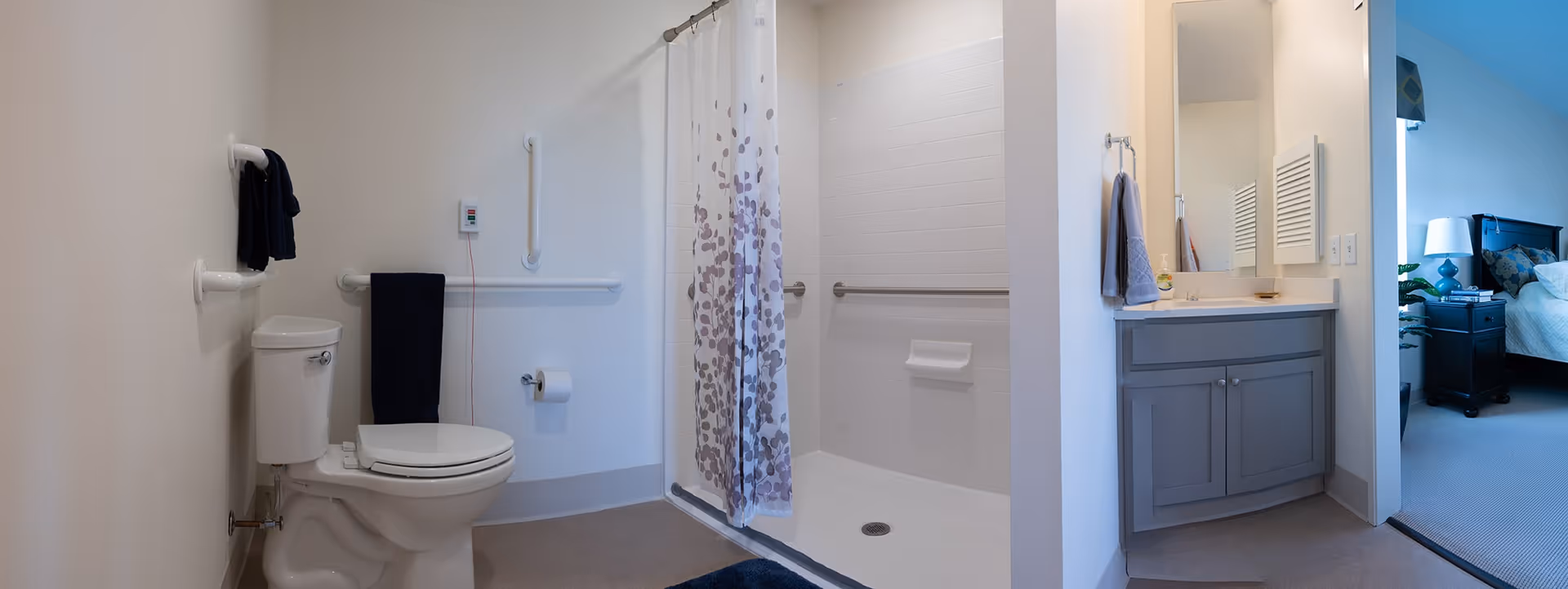 A clean and accessible bathroom with a white toilet, grab bars on the walls, a shower area with a curtain featuring a leaf pattern, and a vanity with a sink and mirror. The bathroom opens into a bedroom with a bed and nightstand visible.