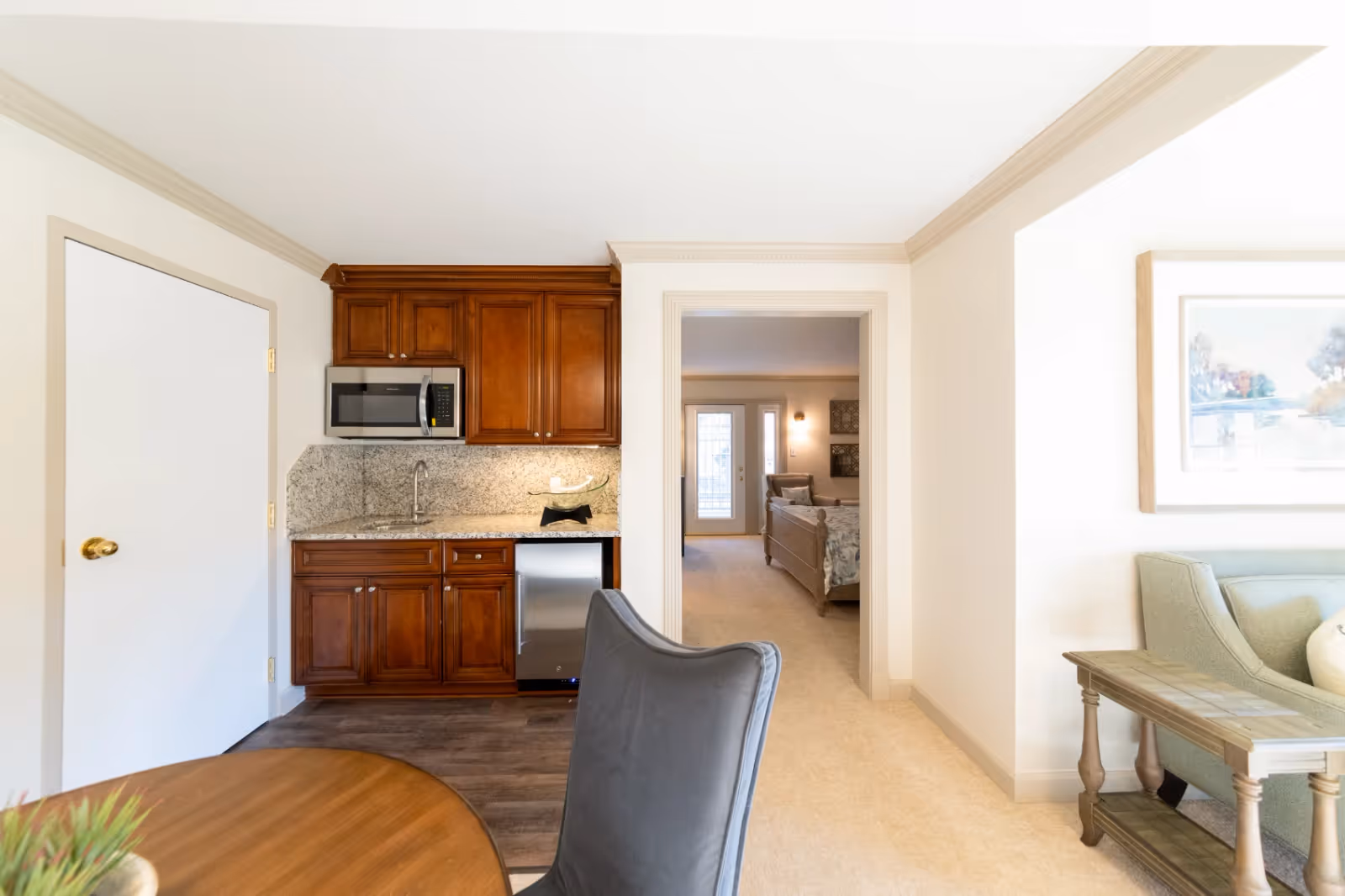 Bright suite interior with a small kitchenette, dining table and seating area leading to a bedroom.
