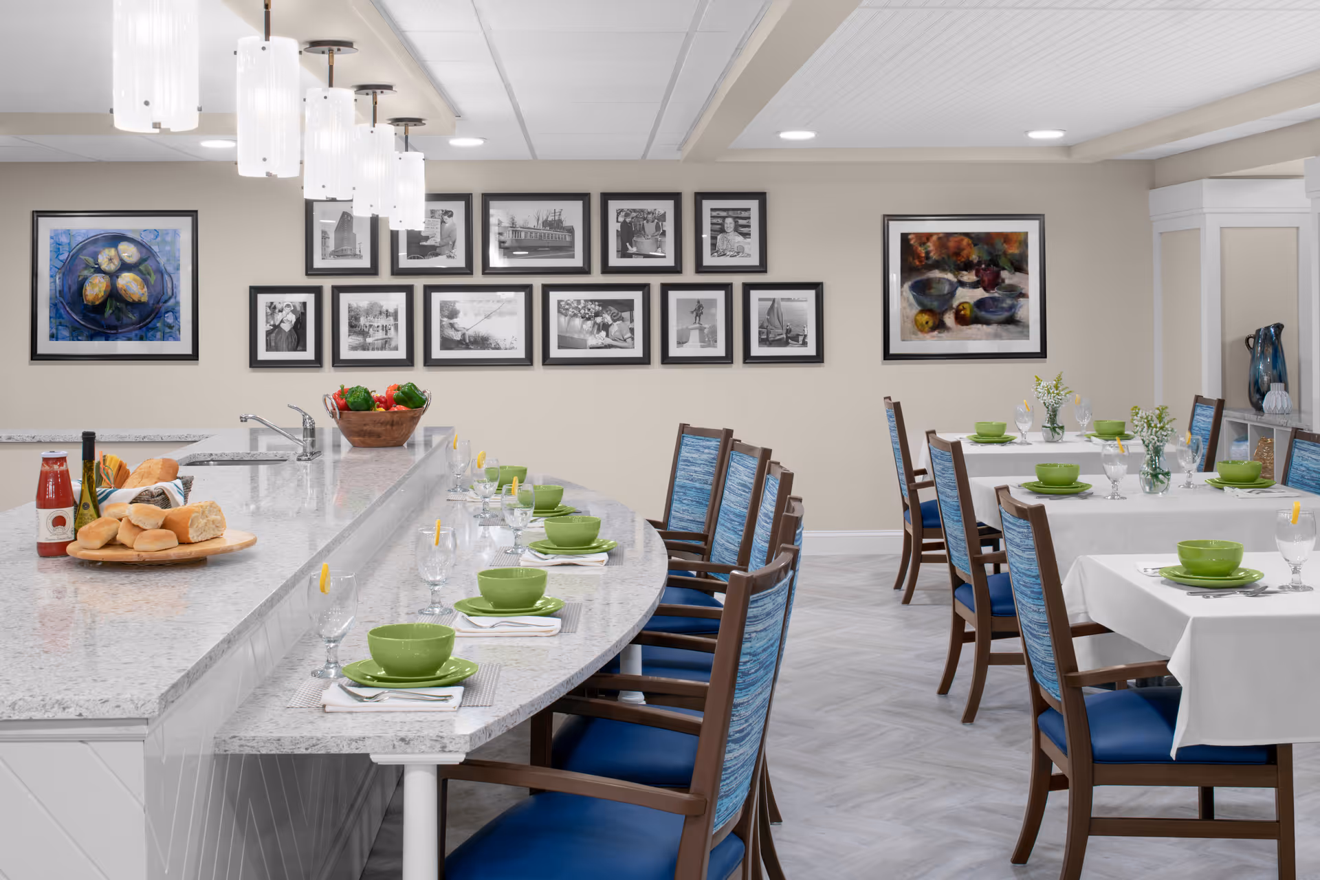 A bright dining area with a curved granite countertop set with green bowls, plates, and glasses with lemon slices. The countertop has a sink, a basket of bread, a bottle of wine, and a bottle of sauce. Behind the countertop are several wooden chairs with blue cushions. In the background, there are tables covered with white tablecloths, also set with green dishware and small flower vases. The walls are decorated with framed black and white photos and colorful paintings. Modern pendant lights hang above the countertop.