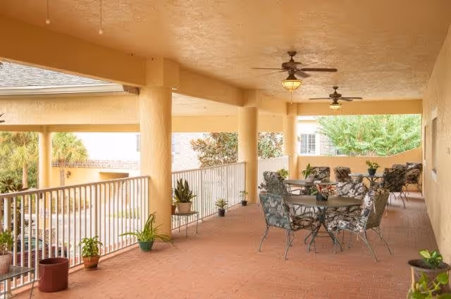 Covered outdoor patio area with several round tables and cushioned chairs with floral patterns. Ceiling fans with lights hang from the ceiling. Potted plants are placed along the railing and floor. The area overlooks greenery and part of the building exterior.