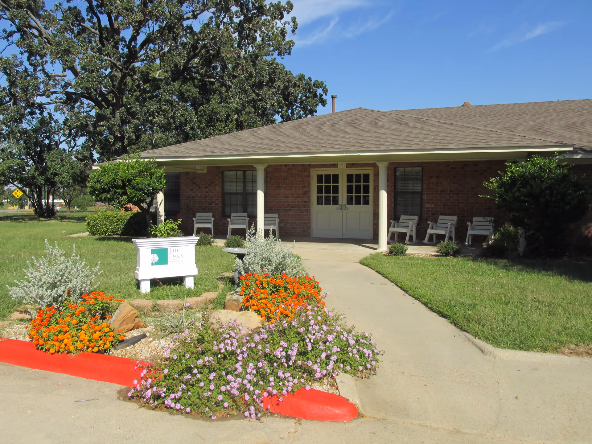 Single-story brick building with a covered porch featuring several white chairs. A concrete walkway leads to double doors at the entrance. In front of the building is a landscaped area with colorful flowers and a white sign that reads 'The Oaks'. Large trees and a clear blue sky are visible in the background.
