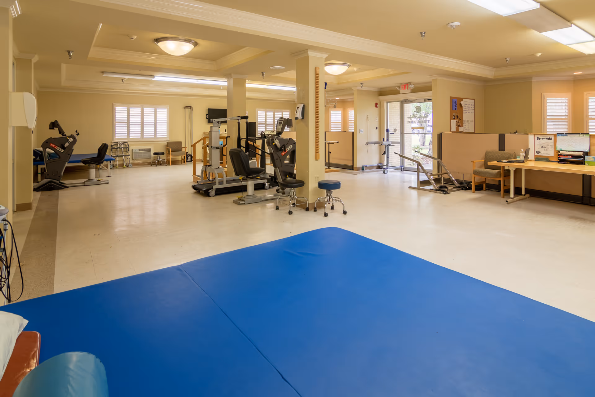 A spacious rehabilitation or physical therapy room in a skilled nursing facility with exercise equipment, chairs, a blue padded mat on the floor, and a desk area near the entrance door. The room has beige walls, large windows with blinds, and bright overhead lighting.