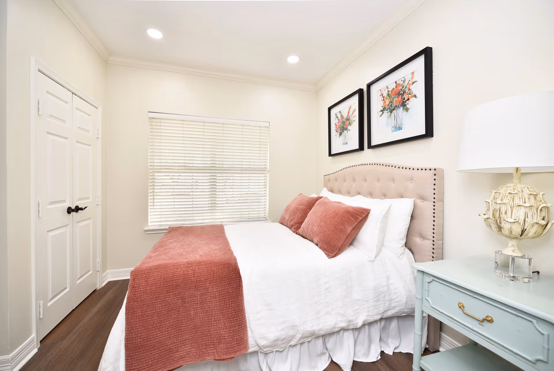 A bright bedroom with a neatly made bed featuring a tufted beige headboard, white bedding, and pinkish-red pillows and throw blanket. There are two framed floral paintings on the wall above the bed, a window with closed blinds, a light blue nightstand with a decorative lamp, and a closed white double-door closet.