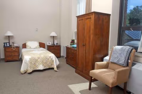 A furnished senior living bedroom with a bed, armoire, chair, and nightstands by a window.