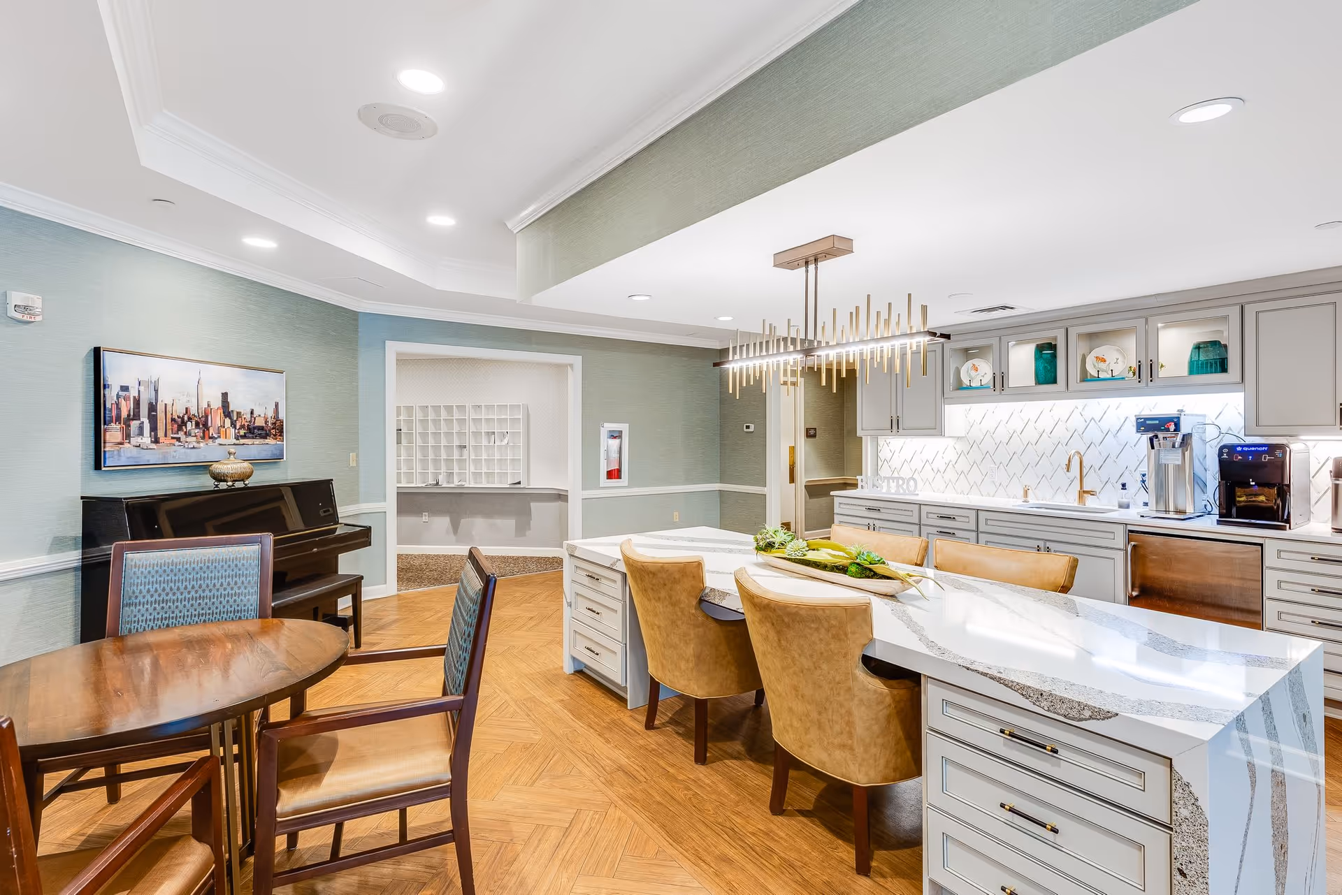 A bright and modern communal kitchen and dining area in a senior living facility. The space features a large marble island with four tan leather chairs, white cabinetry with gold handles, a coffee machine, and a dishwasher. There is a round wooden table with four chairs in the foreground and a piano against the wall with a framed cityscape artwork above it. The room has wood-patterned flooring and recessed ceiling lights.
