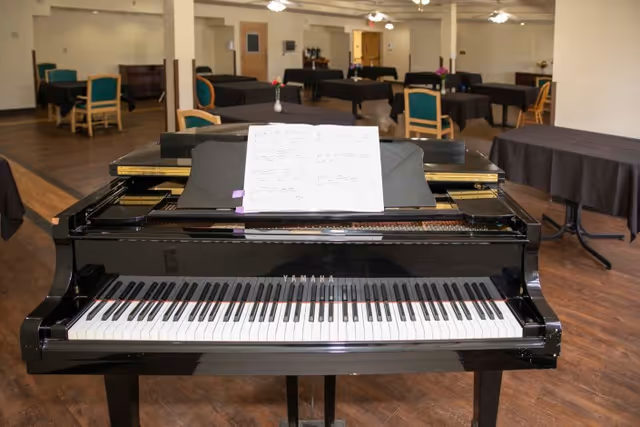 A black Yamaha grand piano with sheet music on the stand is positioned in a spacious room with wooden floors. In the background, there are multiple tables covered with black tablecloths and green cushioned chairs arranged around them. The room appears to be a common area or lounge with neutral-colored walls and ceiling fans.
