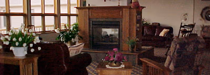 Cozy communal living room with a central fireplace, upholstered seating, coffee tables and potted plants near large windows.