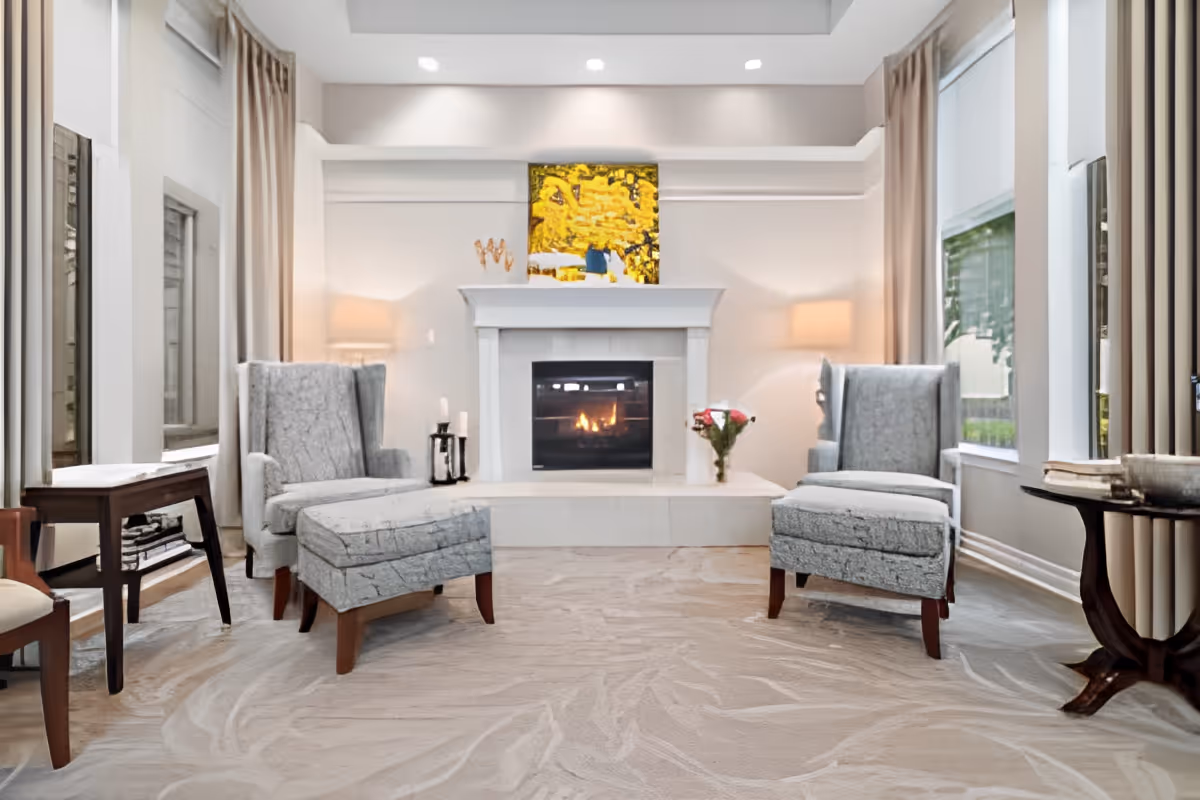 A cozy living room with two gray upholstered armchairs and matching ottomans facing a white fireplace with a fire burning inside. The room has large windows with beige curtains, a beige patterned carpet, a small side table with a vase of flowers, and soft lighting from two lamps on either side of the fireplace.