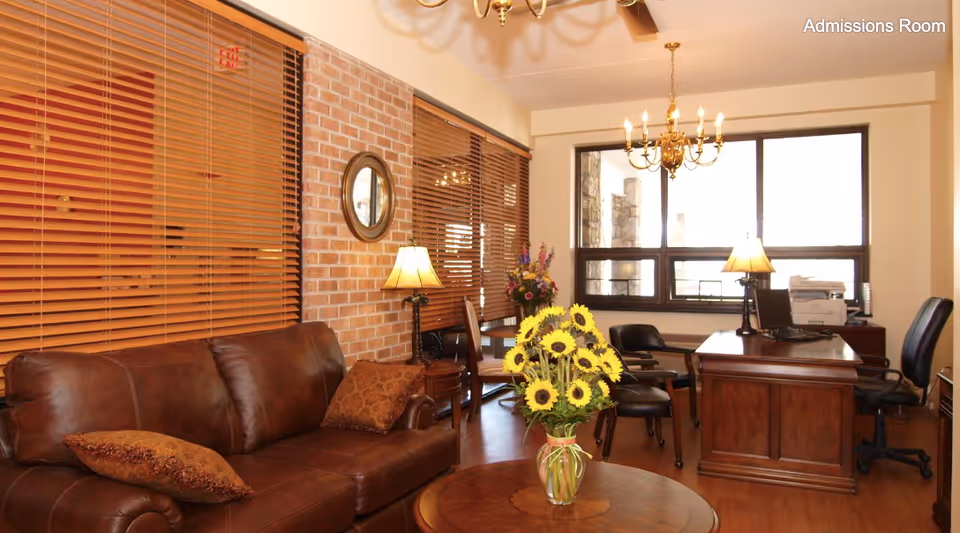 A warmly lit admissions room featuring a brown leather sofa with decorative pillows, a round wooden coffee table with a vase of sunflowers, a wooden desk with office chairs, lamps, and a large window with wooden blinds. The room has brick and cream-colored walls with a chandelier hanging from the ceiling.
