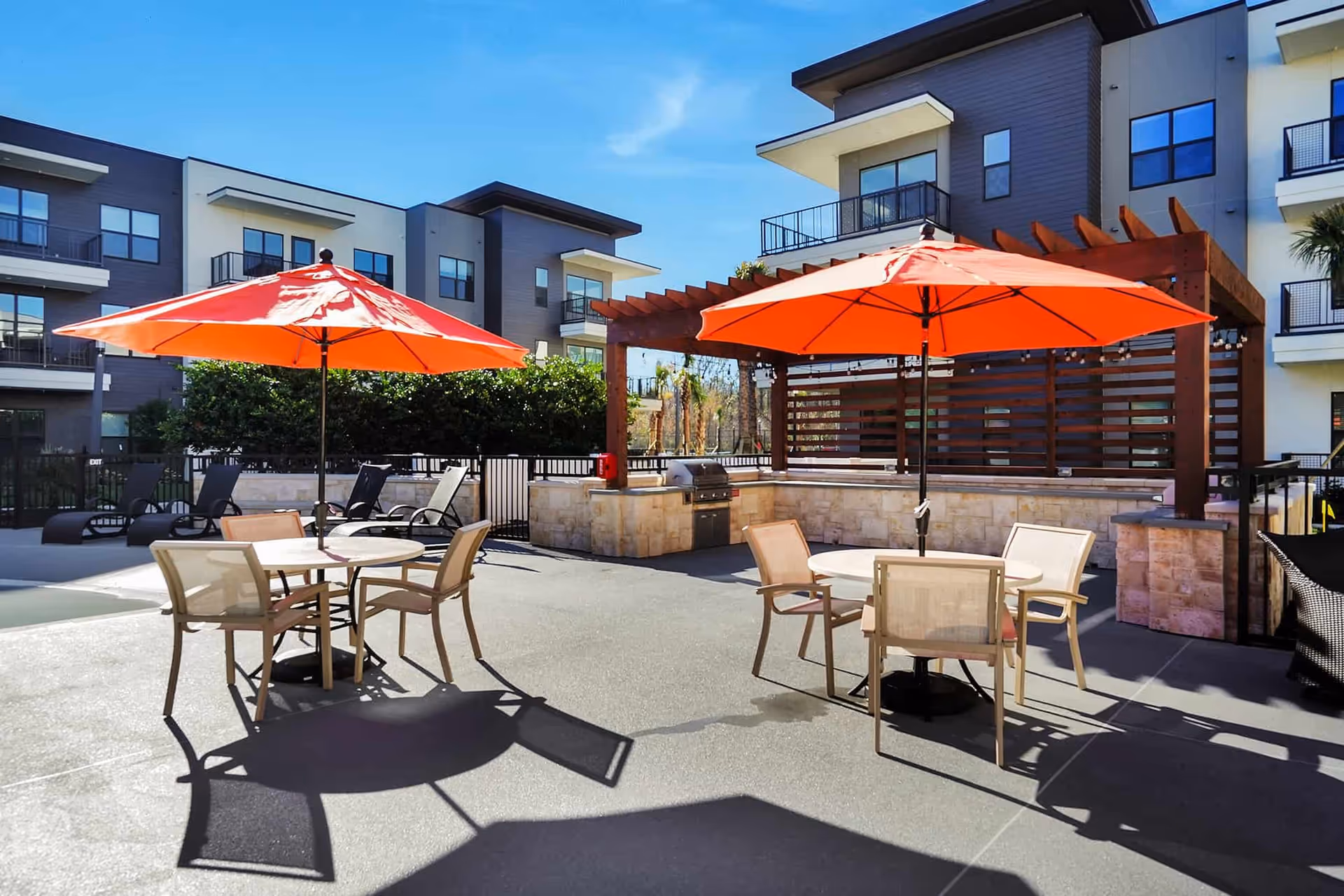 Outdoor patio area at Discovery Village Deerwood Independent Living featuring two round tables each with four chairs and large orange umbrellas providing shade. In the background, there is a built-in grill under a wooden pergola and modern apartment-style buildings with balconies.