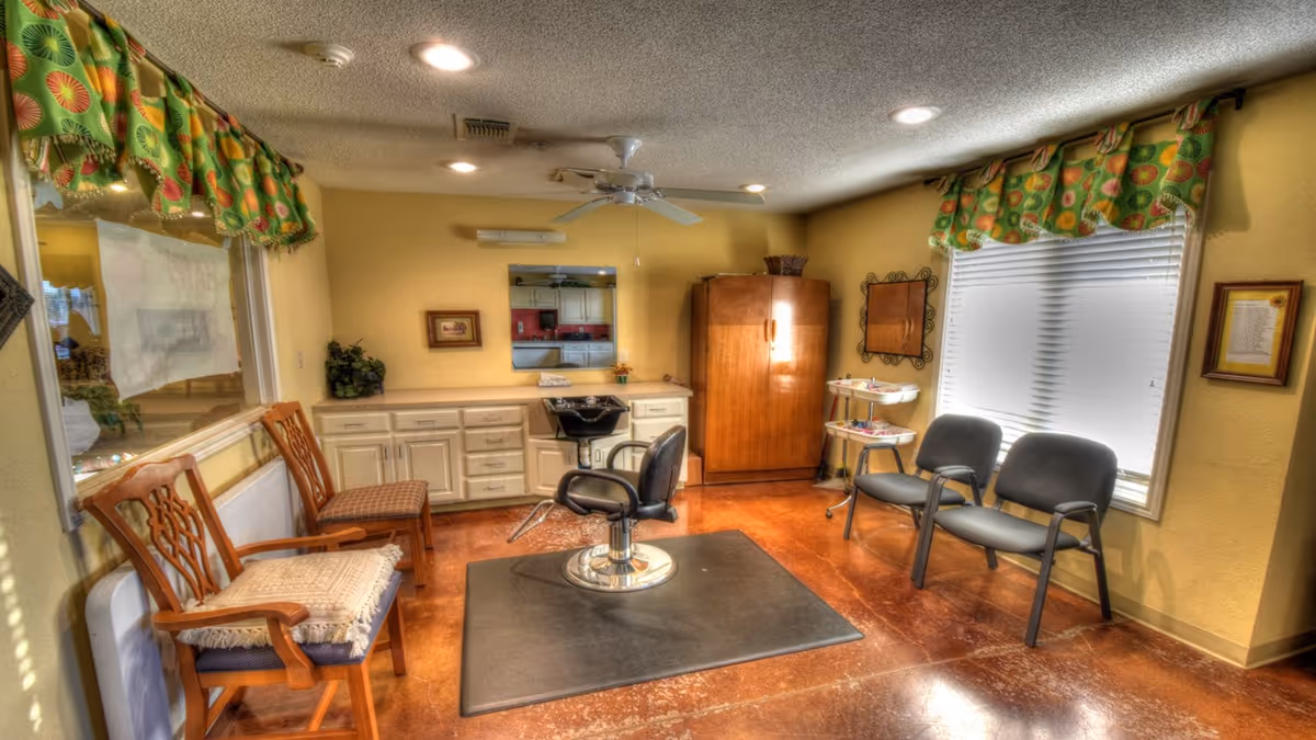 Small salon-style room with a central styling chair and sink, waiting chairs, cabinets and windows with colorful valances.