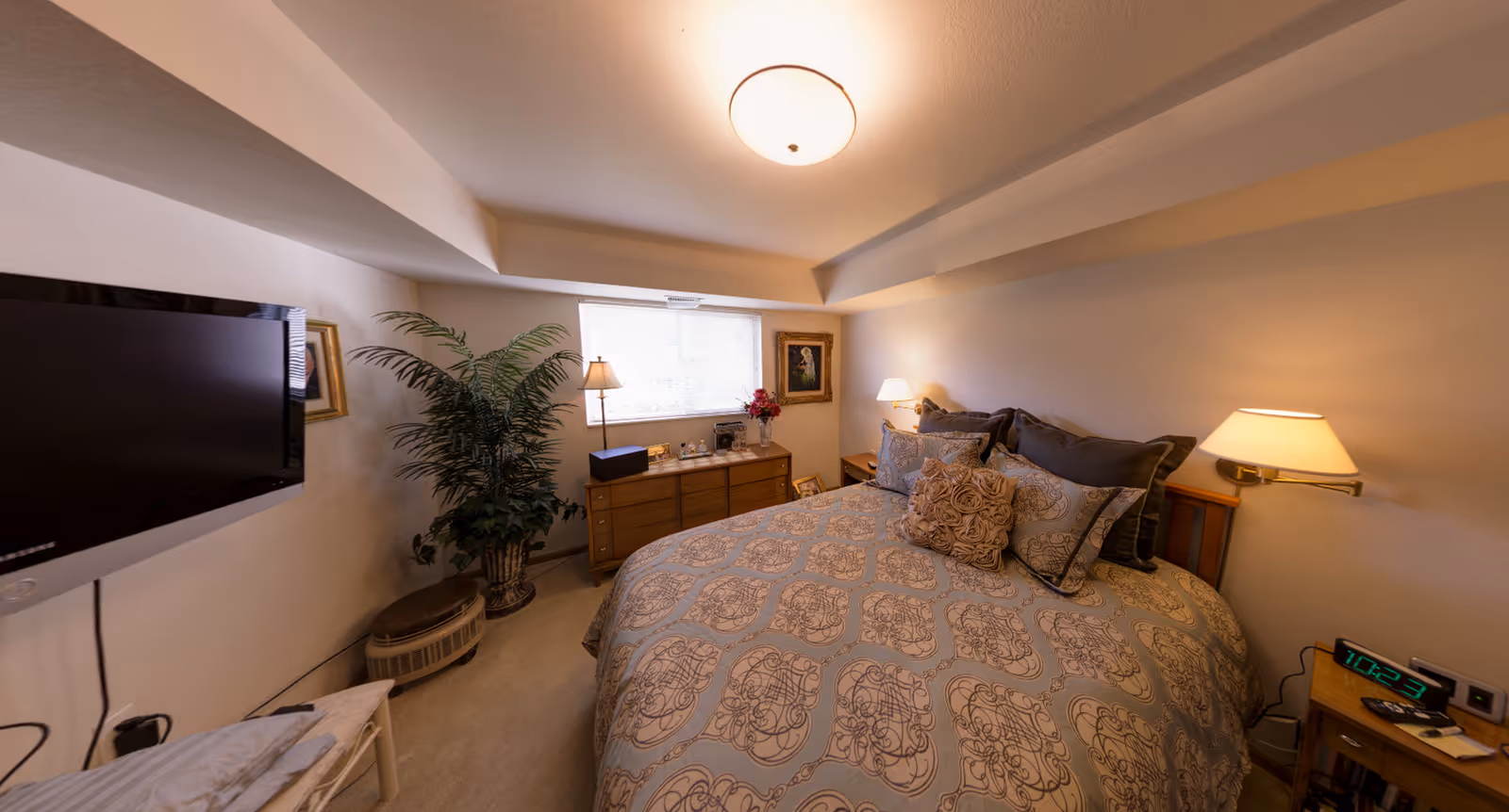 Cozy furnished bedroom with a patterned bed, bedside lamps, a wall-mounted TV, dresser and potted plant.