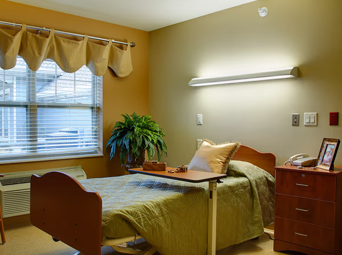 A neatly made single bed with a green quilt and a beige pillow in a senior living facility room. A wooden bedside table with a telephone and a framed photo is next to the bed. A small overbed table is positioned over the bed. There is a large window with blinds and a valance, and a green potted plant sits on a small table near the window. A long wall-mounted light fixture illuminates the room.