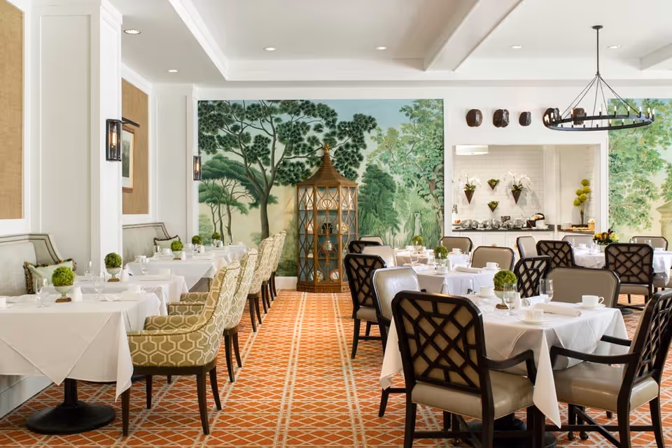 A bright and elegant dining room with tables covered in white tablecloths, set with glassware and small green topiary centerpieces. The room features patterned carpet flooring, cushioned chairs with different upholstery designs, and a large mural of trees and greenery on the far wall. There is a decorative wooden cabinet and a buffet area with plants and serving dishes in the background.