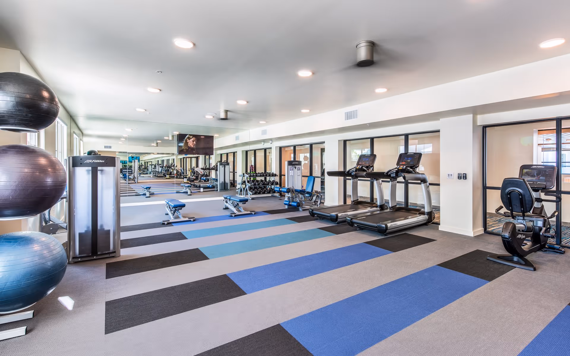 A spacious fitness room with exercise equipment including treadmills, stationary bikes, weight benches, dumbbells, and large exercise balls. The room has a colorful striped carpet and large mirrors on one wall reflecting the equipment.