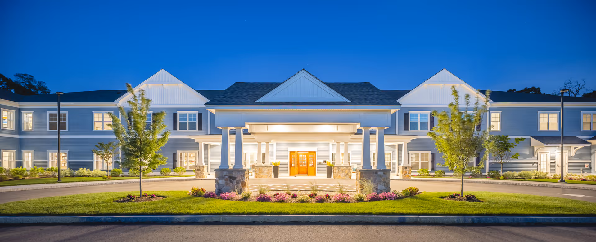 Front exterior view of The Branches of Marlboro building at dusk, with well-lit entrance, manicured lawn, small trees, and flower beds.