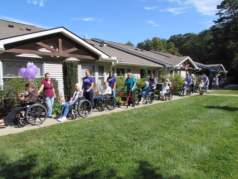 A group of elderly individuals, many in wheelchairs, accompanied by caregivers, are outdoors on a sunny day walking along a sidewalk next to a single-story building with beige siding and brown trim. The scene is set in a green grassy area with trees in the background under a clear blue sky.