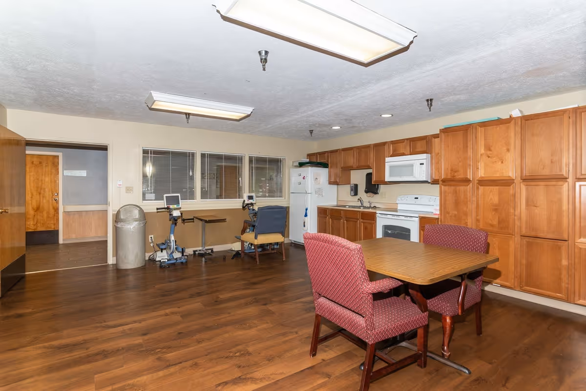 Interior view of a senior living facility kitchen and dining area with wooden cabinets, a white refrigerator, microwave, and stove. There is a wooden table with two upholstered chairs in the foreground. In the background, there are two exercise bikes and a small table with a chair near a window. The floor is wooden, and the ceiling has fluorescent lights.