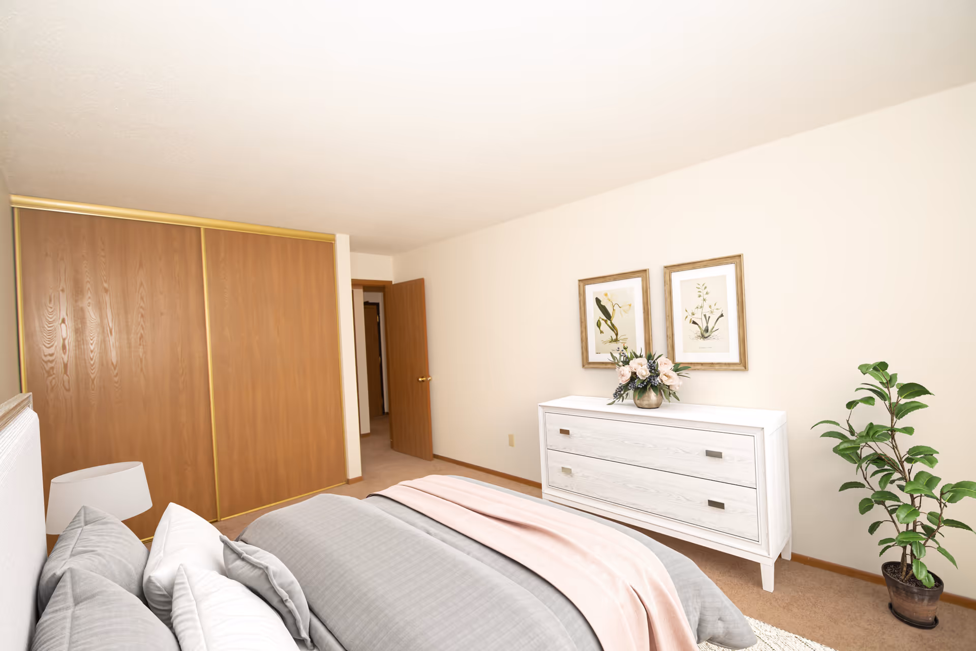 A bedroom with a bed featuring gray and white bedding and a pink throw blanket. There is a white dresser with a flower arrangement on top, two framed botanical prints on the wall above the dresser, a potted plant on the floor, a wooden closet with sliding doors, and an open wooden door leading to another room.