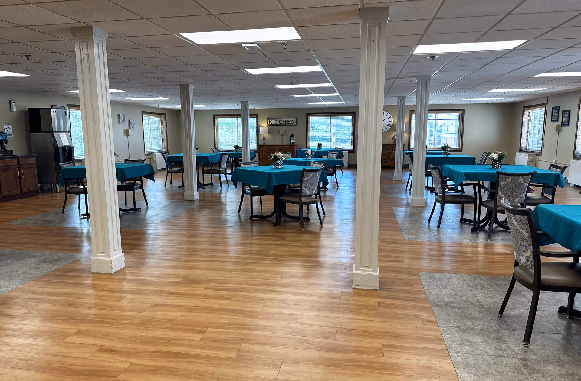 Large bright dining room with multiple round tables covered in teal tablecloths, chairs, support columns, and wood flooring.