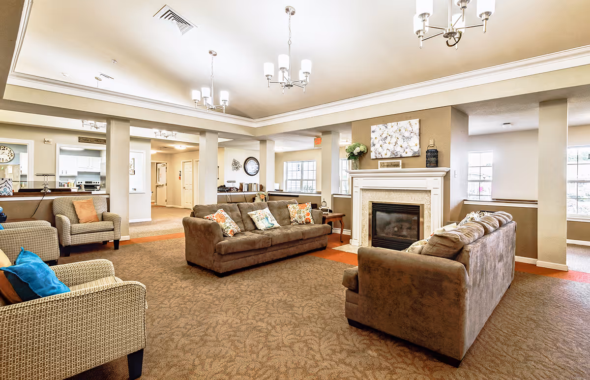 A spacious and well-lit living room area in a senior living facility named Walker Place, featuring two brown sofas with decorative pillows, two patterned armchairs with colorful cushions, a fireplace with a mantel decorated with flowers and artwork, beige walls, carpeted floor, and multiple ceiling light fixtures.
