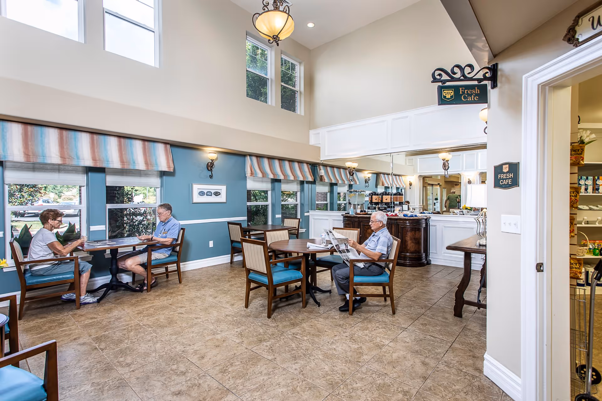 A bright and spacious cafe area inside a senior living facility with large windows and high ceilings. There are several tables and chairs, with two elderly people sitting at one table and another elderly person sitting alone at a different table reading a newspaper. The walls are painted blue with white trim, and there is a sign that reads 'Fresh Cafe' hanging above the entrance to a small serving area.