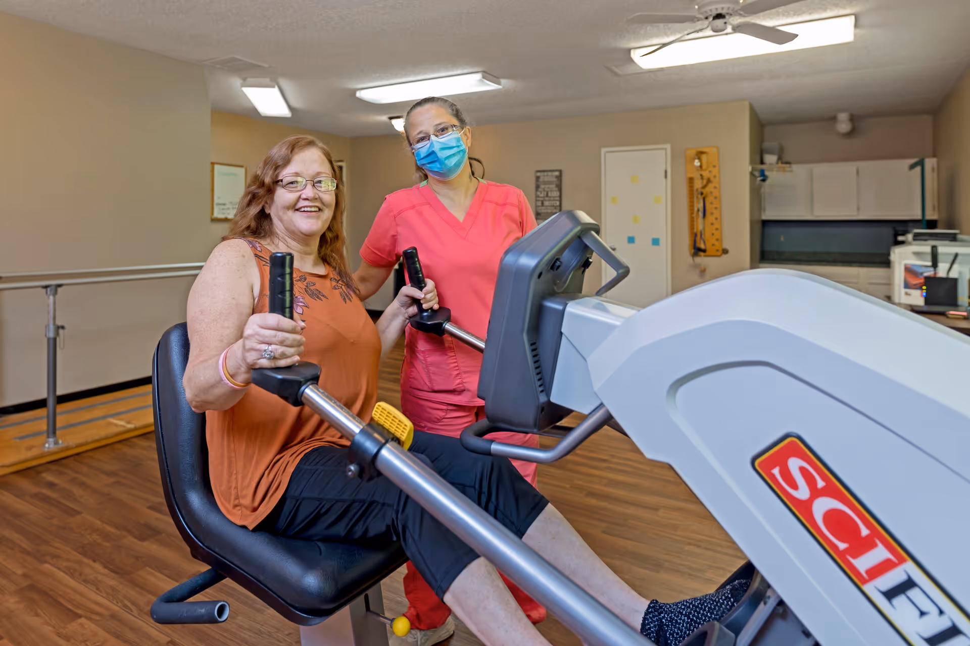 A woman in an orange sleeveless top and black pants is seated on a rowing exercise machine, smiling at the camera. A healthcare worker in pink scrubs and a blue face mask stands behind her, providing support. The room has wooden flooring, handrails along the wall, and medical or rehabilitation equipment in the background.