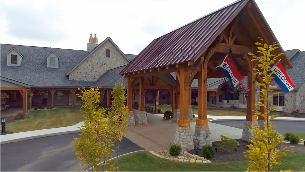Front entrance of a senior living facility with a wooden covered porte-cochere, stone exterior, landscaping, and welcome flags.