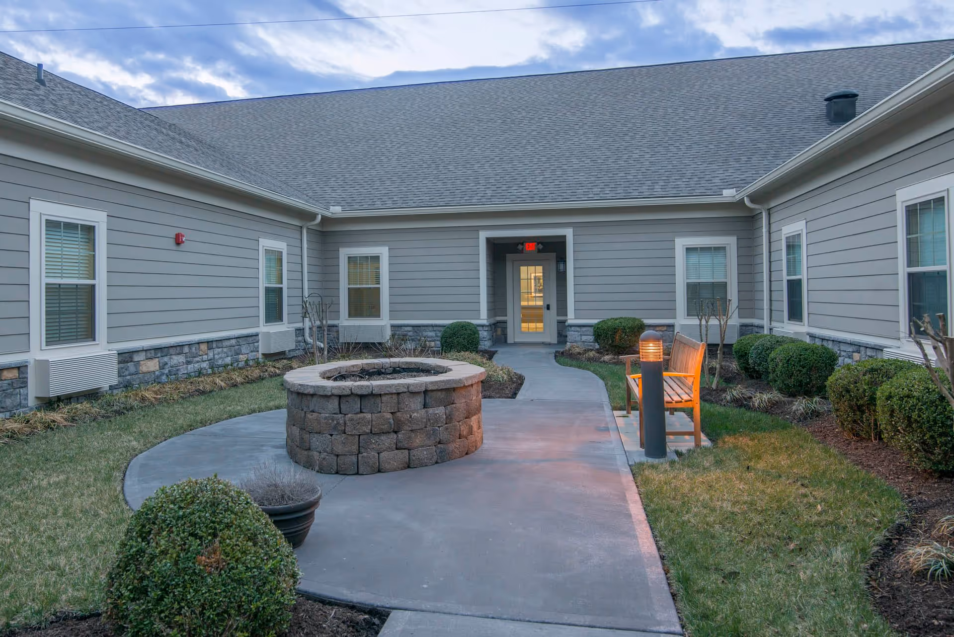 Courtyard of a senior living facility with a circular stone fire pit, paved walkway, bench, shrubs, and an entry door to the building.