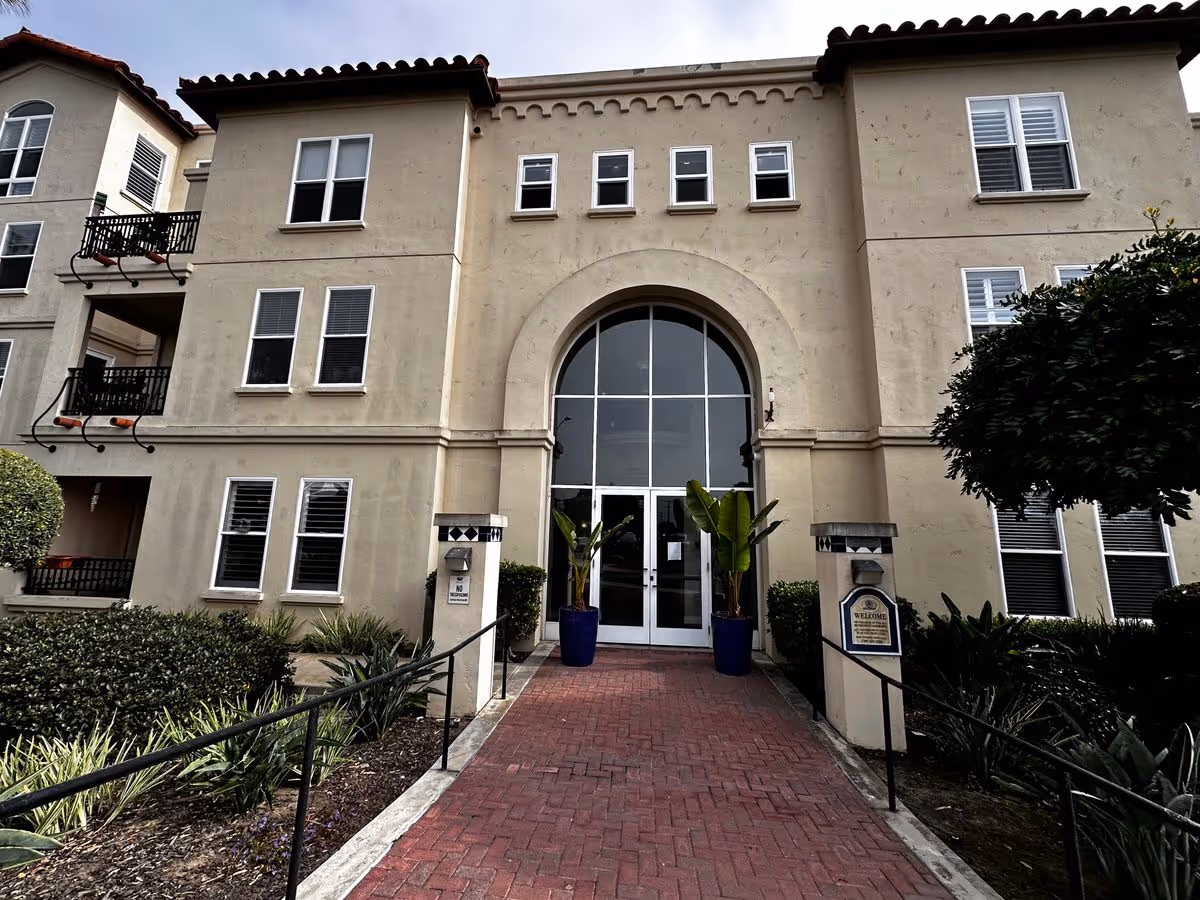 Front entrance of a three-story stucco building with an arched glass doorway, potted plants, and a red brick walkway.